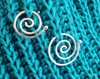 May include: Two silver-toned metal spiral pins with rounded ends, each featuring a coiled wire design. These circular fasteners are positioned against a background of vibrant teal knitted fabric with a visible textured stitch pattern.