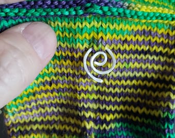 May include: A silver spiral stitch marker rests on a knitted fabric. The fabric features a vibrant mix of green, yellow, and purple yarns. The stitch marker is a simple, elegant design.