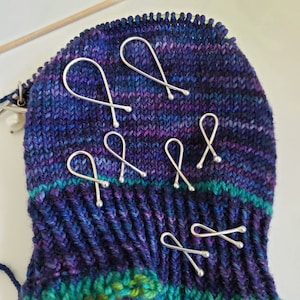 May include: A collection of silver stitch markers in a bow shape, resting on a vibrant, hand-knitted piece of fabric. The fabric features a mix of blue, purple, and green hues, with visible knitting needles in the background. These markers are designed for knitting projects.