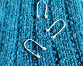 May include: Three silver metal stitch markers in the shape of a U, on a blue knitted fabric background.