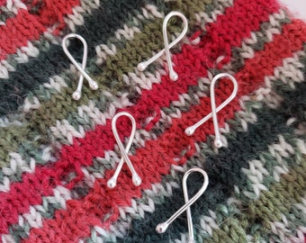 May include: Five silver stitch markers with a looped design and a small bead at the end, resting on a colorful knitted fabric. The fabric features stripes of red, green, white, and black.