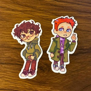 May include: Two cartoon stickers featuring characters with unique styles. One has curly brown hair, glasses, and a green hoodie. The other has red hair, blue eyes, and a green coat, giving a peace sign. Both are wearing sneakers.