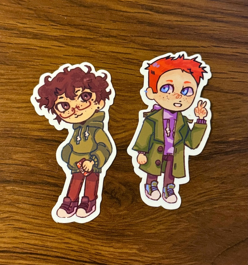 May include: Two cartoon stickers featuring characters with unique styles. One has curly brown hair, glasses, and a green hoodie. The other has red hair, blue eyes, and a green coat, giving a peace sign. Both are wearing sneakers.
