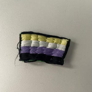 Op de afbeelding: Een klein geborduurd pleister met de kleuren van de non-binaire pride-vlag. De vlag is geel, wit, paars en zwart. De pleister is omgeven door een zwarte rand.