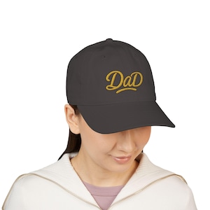 Op de afbeelding: Een donker antracietgrijze honkbalpet met het woord "DaD" geborduurd in een goudgeel cursief lettertype op het voorpaneel. De pet heeft een gestructureerde kroon, een gebogen klep en een effen ontwerp met bijpassende stiksels over het geheel.