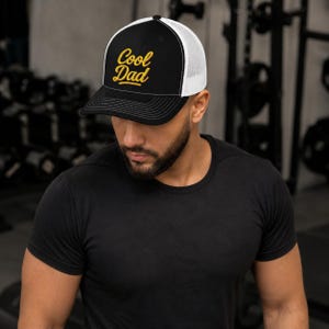 Könnte beinhalten: Eine schwarz-weiße Trucker-Cap mit dem in gelber Schreibschrift gestickten Text Cool Dad auf der Vorderseite. Die Kappe hat eine strukturierte schwarze Front, eine weiße Netzrückseite und einen schwarzen Schirm mit weißen Ziernähten am Rand.