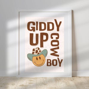 May include: A white framed print with a brown background and the text "Giddy Up Cow Boy" in brown letters. A smiling cartoon face wearing a blue and brown cowboy hat is in the center of the print.