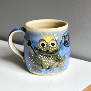 May include: A ceramic mug with a light blue glaze featuring a hand-painted green frog wearing a yellow crown. The frog has white polka dots and is holding a black stick. A speech bubble next to the frog contains the text, "POOF You are danger."
