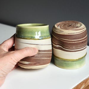 May include: Two handmade ceramic tumblers. One is held by a hand. The tumblers feature a green rim, brown and white horizontal stripes, and a cylindrical shape. The other tumbler is upside down.