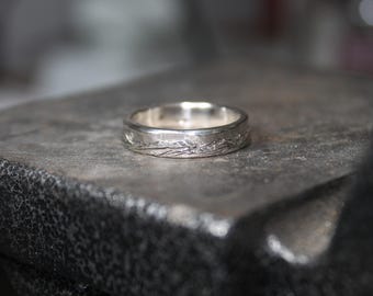 May include: A silver band with a textured design. The band is smooth and shiny, with a subtle pattern of lines and curves.