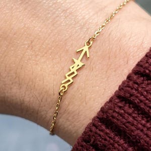 May include: A gold-toned chain bracelet featuring a central charm shaped like an arrow with a jagged, lightning-bolt-style shaft. The delicate metal chain connects to both ends of the arrow charm, creating a continuous link design around the wrist.