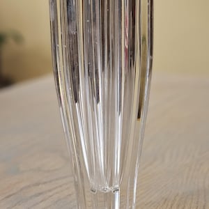 Pu&ograve; includere: Vaso di vetro trasparente con design a coste verticali. Il vaso ha una parte superiore svasata e una base geometrica. Il vetro trasparente riflette la luce, creando ombre. Il vaso &egrave; probabilmente per scopi decorativi.