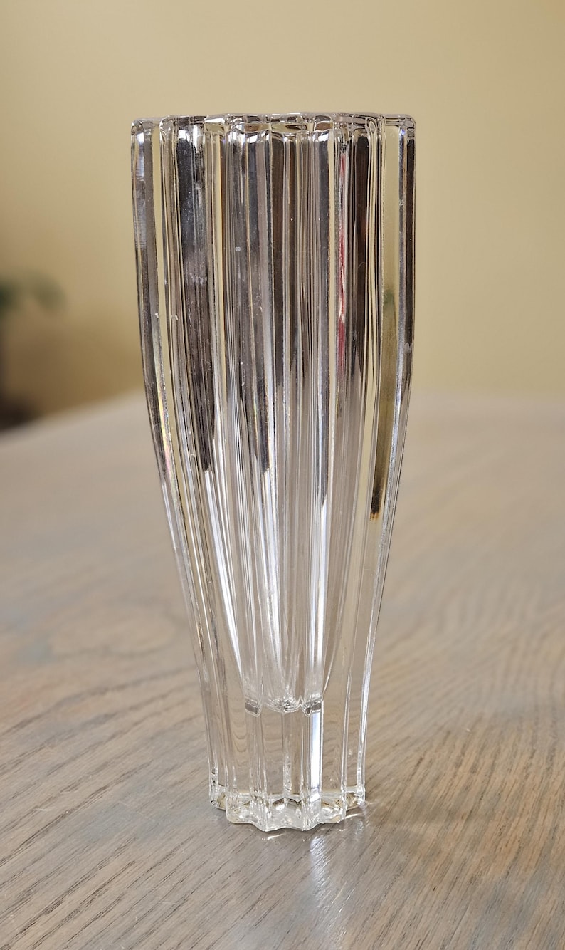 Pu&ograve; includere: Vaso di vetro trasparente con design a coste verticali. Il vaso ha una parte superiore svasata e una base geometrica. Il vetro trasparente riflette la luce, creando ombre. Il vaso &egrave; probabilmente per scopi decorativi.