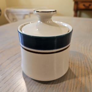 May include: A ceramic sugar bowl with a lid. The bowl is off-white with a dark blue band and a thin brown stripe. The lid and handle are off-white with a thin brown trim. The bowl is cylindrical and sits on a wooden table.