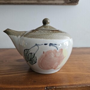 May include: A ceramic teapot with a spout and a lid. The teapot is white with a brown and tan rim and lid. It features a hand-painted design of pink and yellow flowers with green leaves. The teapot is sitting on a wooden surface.