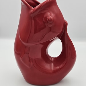 May include: A glossy, deep red ceramic pitcher shaped like a stylized fish. The vessel features a wide, open mouth at the top, a smooth, curved body, and a large, circular cutout near the base that forms the handle. The surface has a reflective finish.