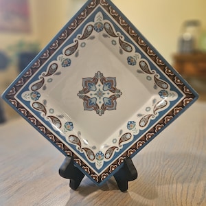 May include: Square ceramic serving dish with a white center and a decorative border in shades of blue and brown. The dish features a central floral design and paisley motifs, displayed on a black stand.