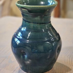 May include: A handcrafted, dark teal ceramic vase with a glossy finish. The vase has a rounded body, a flared neck, and subtle textured rings around the middle. Two small, circular indentations are visible on the sides.