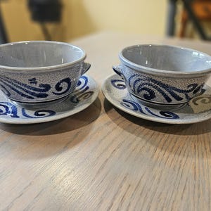 May include: Two matching gray ceramic soup bowls with handles, each resting on a matching saucer. The bowls and saucers feature a blue wave and swirl design. The set is displayed on a light wood surface.