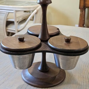 May include: A wooden condiment server with three stainless steel bowls. Each bowl has a wooden lid with a small knob. The server is dark brown and sits on a round base. The design is mid-century modern.