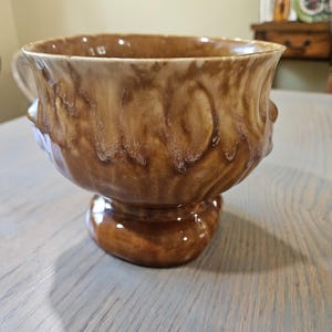 May include: A brown ceramic pedestal bowl with a handle. The bowl has a textured, glazed surface with a dripped design. The bowl is on a pedestal base. The interior of the bowl is a lighter brown color.