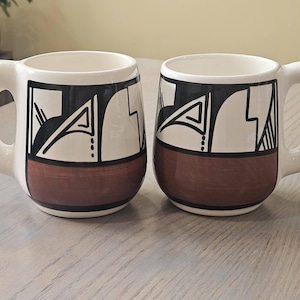 May include: Two white ceramic mugs with a brown base and geometric black and white designs. Each mug has a large, rounded handle. The mugs are displayed on a light gray surface.