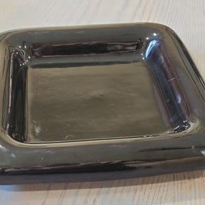 May include: A glossy, black square dish with rounded corners. The dish has a slightly raised edge and a smooth, reflective surface. The dish is empty and sits on a light-colored wooden surface.
