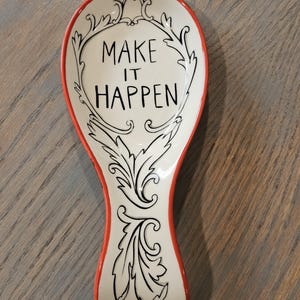 May include: A ceramic spoon rest with a cream-colored surface and a bright orange rim. The center features black hand-lettered text that reads "MAKE IT HAPPEN" surrounded by a decorative black line-art scrollwork pattern that extends down the handle.