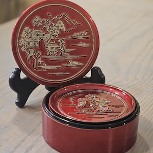 May include: Red and gold decorative coasters with a Japanese landscape design. The coasters are round and feature a detailed relief of a pagoda, trees, and mountains. The set includes a coaster holder.