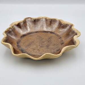 May include: A shallow ceramic bowl with a wavy, scalloped rim. The interior features a metallic bronze and dark brown glaze with a textured, mottled finish, while the exterior rim displays a matte, light beige, unglazed clay appearance.