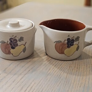 May include: Cream-colored ceramic sugar bowl with lid and creamer set. Each piece features a painted design of a red apple, purple grapes, and a yellow pear. The creamer has a brown interior and a handle.