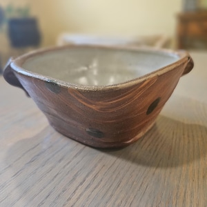 May include: A handmade, square ceramic bowl with a light gray interior and a brown exterior. The bowl has a rustic appearance with dark brown spots and handles on each side. The bowl is sitting on a wooden table.