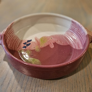 May include: A handmade ceramic bowl with a wide, shallow design. The bowl features a deep burgundy base, a white interior, and a rim with pink and red accents. It has two small handles and is decorated with abstract floral designs.