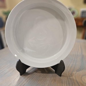 May include: A white ceramic plate with a wide rim, displayed on a dark wooden stand. The plate has a smooth, glossy surface. The stand is a dark brown color and has a simple, functional design. The plate is round and appears to be a serving dish.
