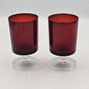 May include: A pair of matching drinking glasses featuring deep red, cylindrical bowls set atop clear, circular glass stems and bases. The glasses have a smooth, uniform finish and a simple, functional design with a wide rim and a sturdy foot.