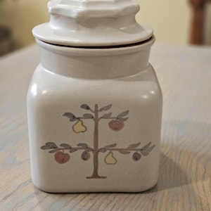 May include: A beige ceramic jar with a lid. The jar has a square shape and a tree design with pears and apples. The lid is a lighter shade of beige and has a square shape. The jar is sitting on a wooden surface.