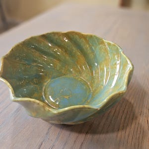May include: A handmade ceramic bowl with a unique, scalloped edge design. The bowl features a blend of teal and gold colors, creating a marbled effect. The interior is a solid teal, and the bowl sits on a wooden surface.