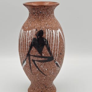 May include: A ceramic vase with a speckled terracotta finish featuring a hand-painted black abstract figure seated in the center. The design includes vertical white brushstrokes surrounding the figure, creating a contrast against the textured brown surface of the vessel.