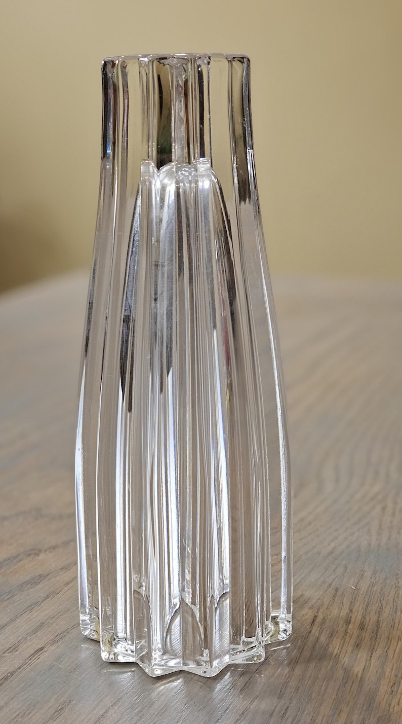 Pu&ograve; includere: Vaso di vetro trasparente con base a forma di stella e design a coste verticali. Il vaso ha una forma alta e sottile con una parte superiore svasata. Il vetro &egrave; trasparente, permettendo alla luce di passare, ed &egrave; posto su una superficie di legno.