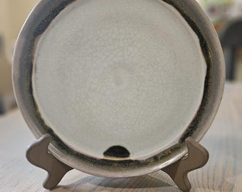May include: A ceramic plate with a light gray center and a dark gray rim. The plate has a crackled texture and is displayed on a wooden stand. The rim has a pinkish hue. The plate is a decorative item.
