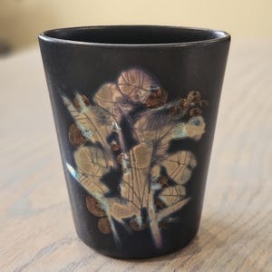 May include: A black ceramic tumbler with a floral design in shades of brown, beige, and blue. The tumbler has a slightly tapered shape and a smooth, glossy finish. The floral pattern is embossed on the surface.