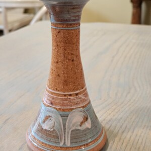 May include: A handmade ceramic candlestick holder with a speckled brown body, accented with blue and white floral designs. The top and base have a light brown and blue trim. The candlestick holder is a decorative home decor item.