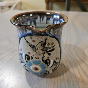 May include: A ceramic creamer with a bird and floral design. The creamer has a brown rim and a light beige body with a black bird illustration. Blue and black floral patterns decorate the base. The creamer has a spout for easy pouring.