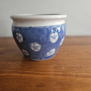 May include: A small, blue ceramic planter with white floral designs. The planter has a rounded shape and a white rim.