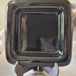 May include: A glossy, square, black dish with rounded corners, displayed on a wooden stand. The dish has a recessed center and a reflective surface. The stand is a dark gray color.