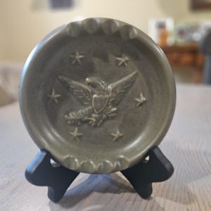 May include: A round, olive-green ashtray with a raised relief of an eagle and stars. The ashtray has a scalloped edge and is displayed on a black stand. The eagle is centered, with stars surrounding it.