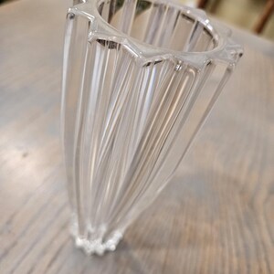 Pu&ograve; includere: Un vaso di vetro trasparente scanalato con un bordo a forma di stella. Il vaso ha una forma svasata e un design strutturato e a coste. La base del vaso presenta un dettaglio decorativo perlato. Il vaso &egrave; appoggiato su una superficie di legno.