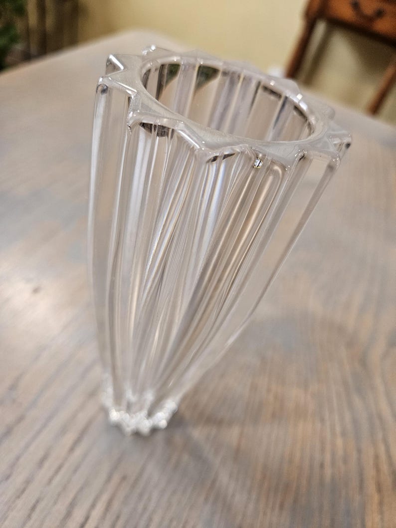 Pu&ograve; includere: Un vaso di vetro trasparente scanalato con un bordo a forma di stella. Il vaso ha una forma svasata e un design strutturato e a coste. La base del vaso presenta un dettaglio decorativo perlato. Il vaso &egrave; appoggiato su una superficie di legno.