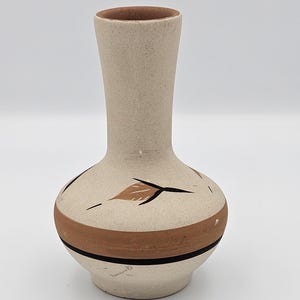 May include: A beige ceramic vase with a narrow neck and a rounded body. The vase features a wide horizontal terracotta band near the base, accented by a thin black line. The upper body is decorated with painted brown leaf motifs and black accents.