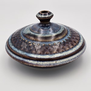 May include: A low-profile ceramic vessel with a wide, rounded body and a small, centered neck opening. The surface features a dark brown base glaze with textured, raised dimples and a reactive blue and white glaze finish around the top.
